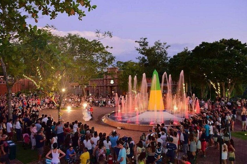Una fuente con múltiples chorros de agua, rodeada de gente, árboles y un edificio al fondo. 3
