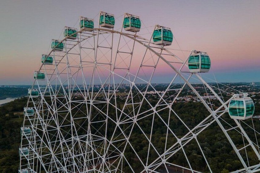 Una noria grande con múltiples cabinas, frente a un cielo al atardecer y un paisaje urbano distante. 1