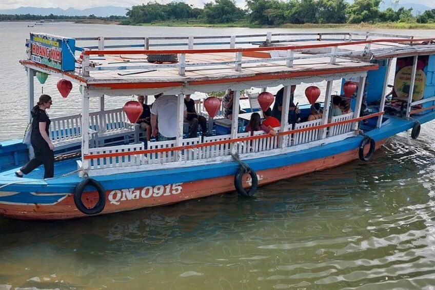 A blue and orange boat with people on board, a person walking beside it, and a river with greenery in the background. 2
