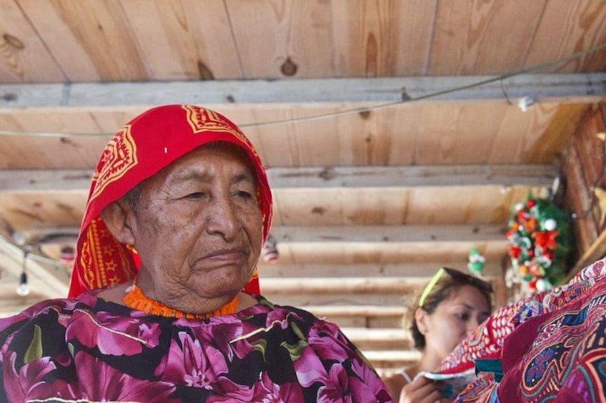 An elderly woman wearing a red patterned headscarf and a floral dress, standing in a wooden structure with a decorated interior. 2