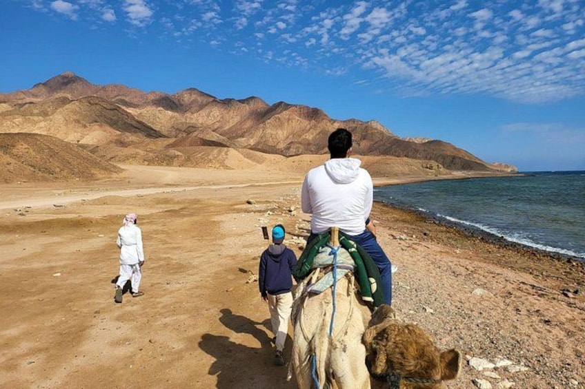A desert landscape with mountains in the background, a camel with riders, and a person walking. 3
