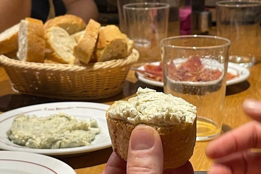 A hand holding a small bread roll with a creamy spread, a basket of bread slices, and a glass of beer on a table. 4