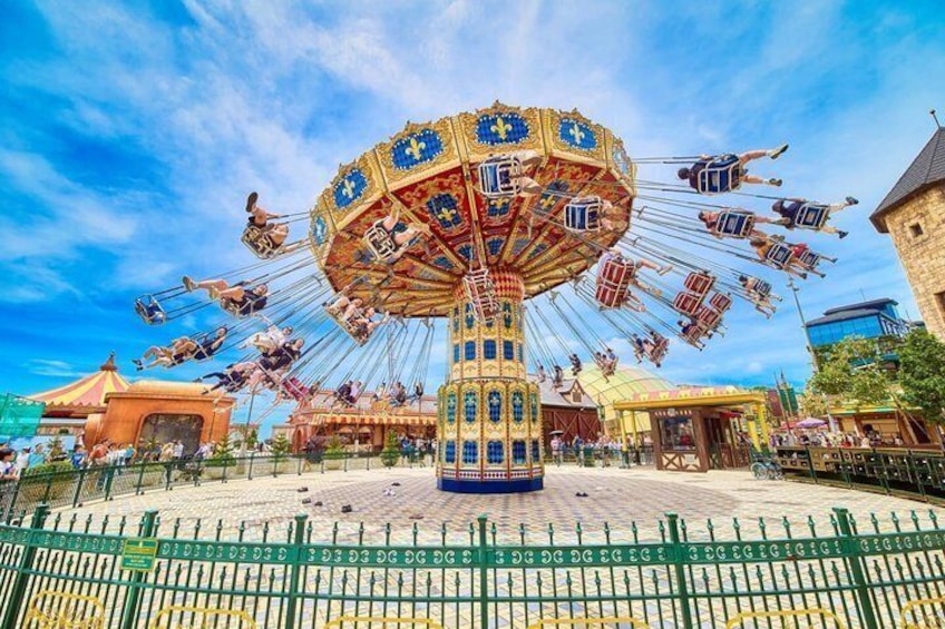 A carousel with riders, surrounded by a fenced area, and buildings in the background. 2