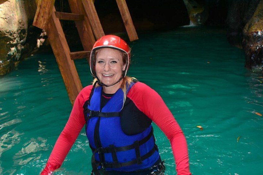 A person in a blue life jacket and red helmet standing in clear water with a wooden structure in the background. 2