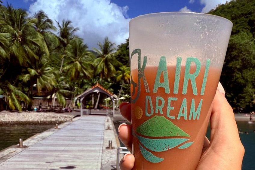 Una mano sosteniendo una taza con la inscripción 'Kairi Dream' y un diseño de hoja de palma, un muelle y una playa tropical con palmeras y una pequeña cabaña al fondo. 4