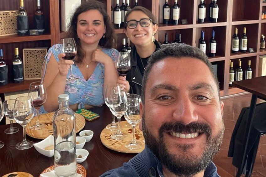 A person holding a wine glass, a table with wine bottles, and a wine rack in the background. 2