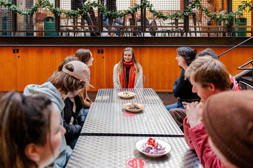 A group of people sitting around a table with food, in an indoor setting with a wooden wall and a railing. 2