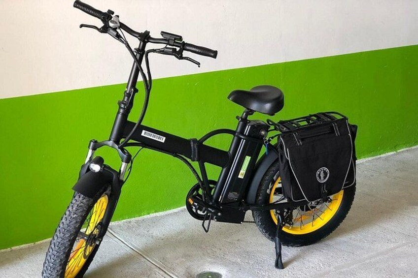 A stationary electric bicycle with a black seat and yellow wheels, parked against a green and white wall. 2