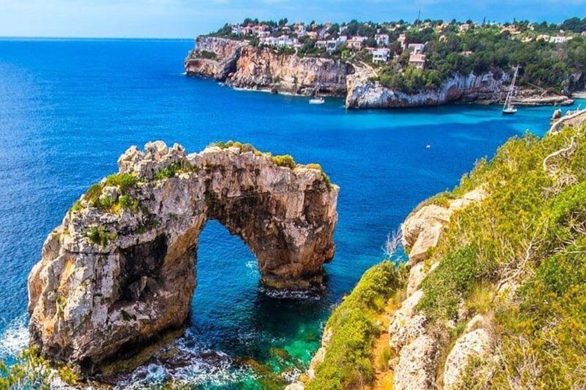 Paisaje costero con un arco natural de roca, mar azul cristalino y un pueblo distante en un acantilado. 5