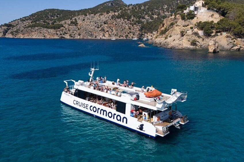A white cruise ship named 'CRUISE CORMORAN' sailing in clear blue waters with a rocky coastline and a hill in the background. 4