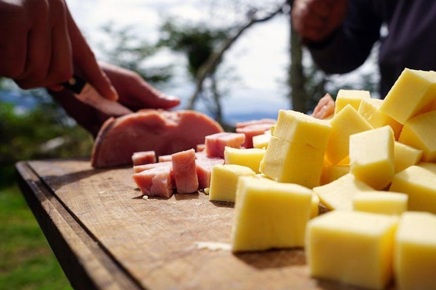 Tagliere di legno con cubetti di formaggio giallo e fette di carne rosa, probabilmente prosciutto, mentre viene preparato all'aperto. 5