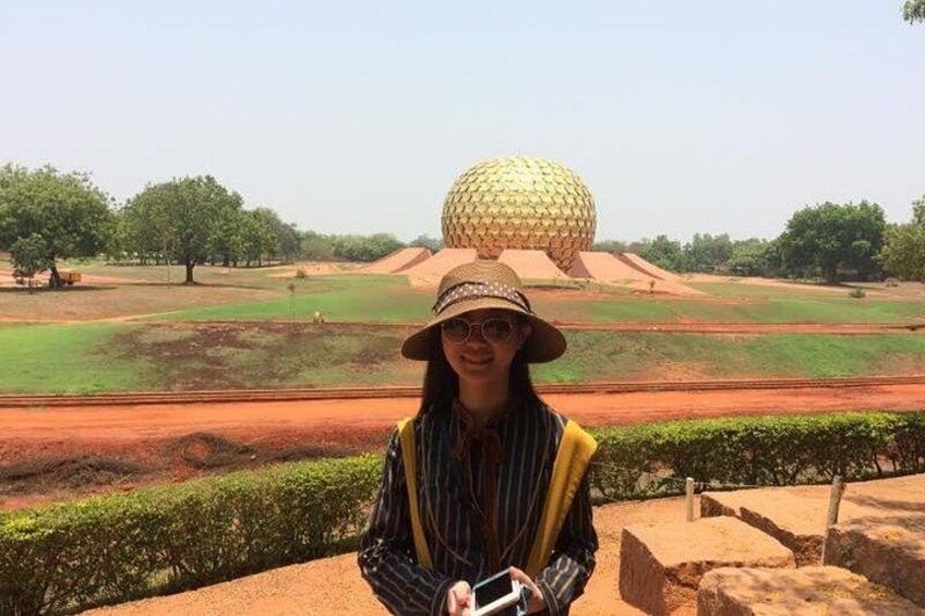 A person standing in front of a large golden sphere structure, with green fields and trees in the background. 1