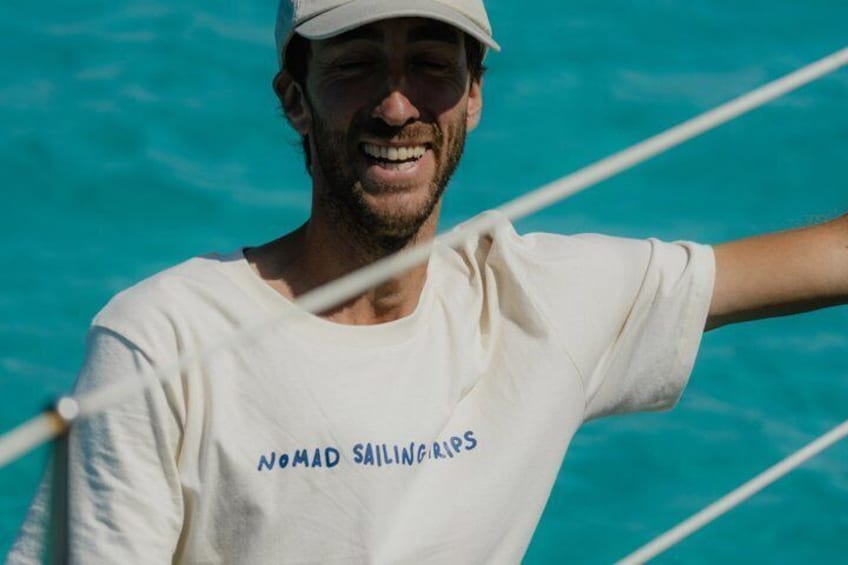 A person wearing a white t-shirt with 'Nomad Sailing Trips' printed on it, standing by a blue body of water. 3
