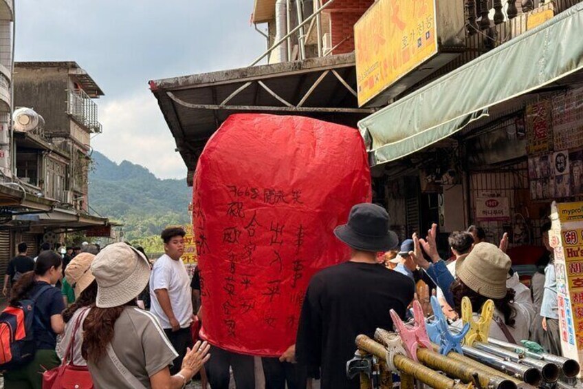A crowd of people carrying a large red lantern with Chinese characters, gathered in a market area with shops and mountains in the background. 3