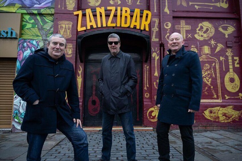 Tres hombres de pie frente a un edificio con un mural y la palabra 'ZANZIBAR' escrita en él. 3