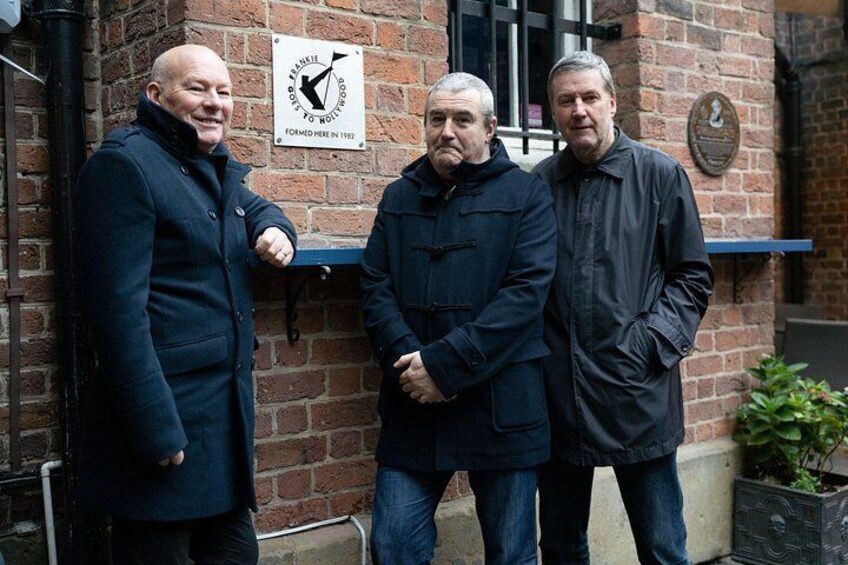 Tres hombres de pie frente a un edificio de ladrillo con un cartel que dice 'BILLINGHAM TO HOLLIS 1992'. 2
