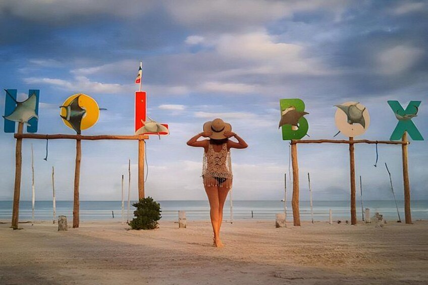 A beach with a woman in a swimsuit and hat, wooden structures with colorful letters, and a flag. 1