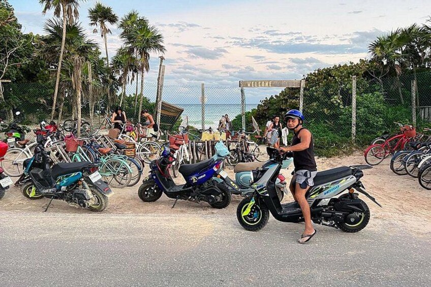 A beachside parking area with multiple scooters and bicycles, palm trees, and a 'WELCOME' sign in the background. 2