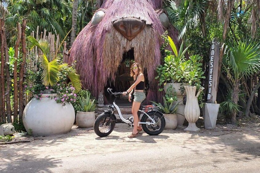 A woman standing next to a white scooter in front of a thatched-roof structure surrounded by tropical plants. 5