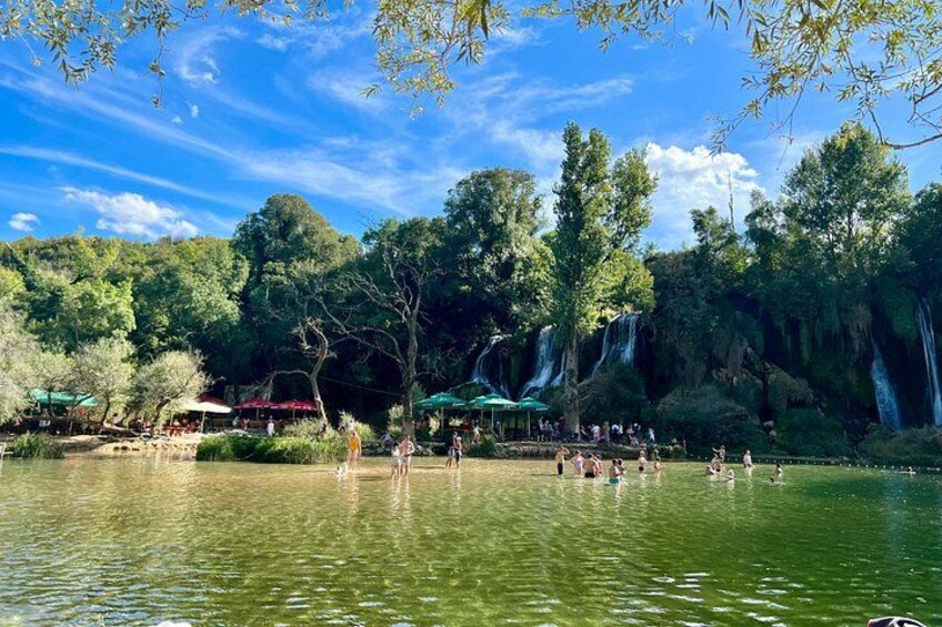 A lake with people swimming, a waterfall, and trees. 2