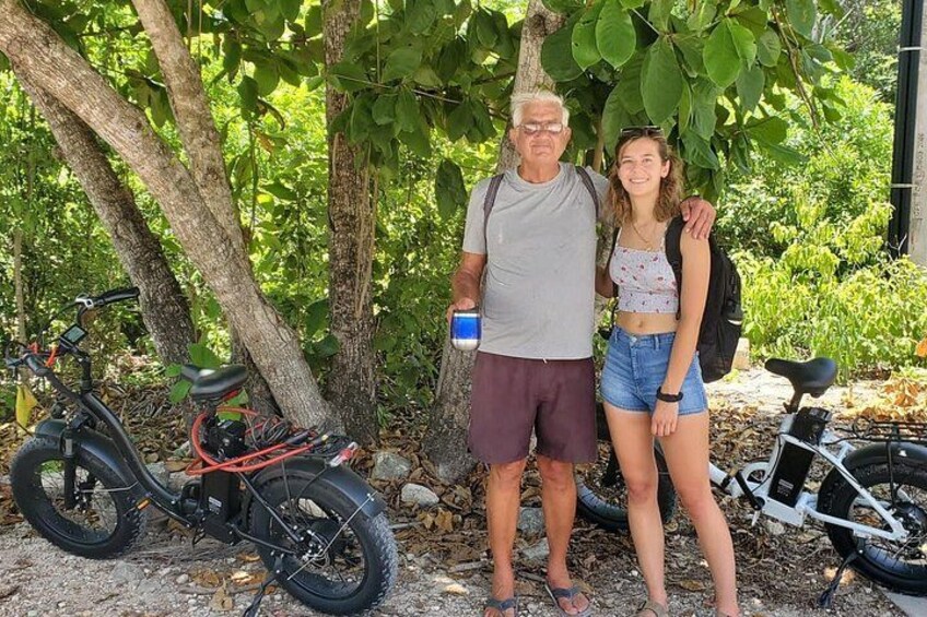 Two people standing by a bicycle and a scooter, with trees and foliage in the background. 2