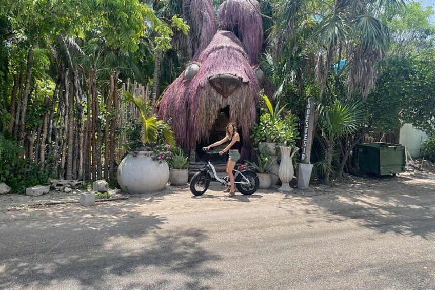 A person sitting on a motorcycle in front of a large thatched structure, surrounded by tropical plants and trees. 4
