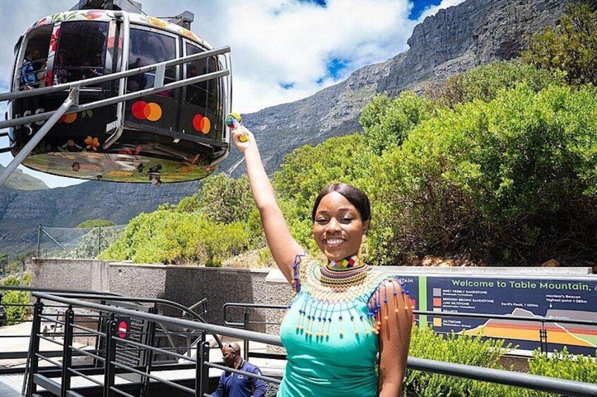 A person standing with a sign in the background, a cable car in the air, and a mountainous landscape. 1