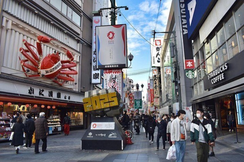 Una concurrida calle urbana con una gran escultura de cangrejo, una cafetería Starbucks y diversas tiendas y escaparates. 3