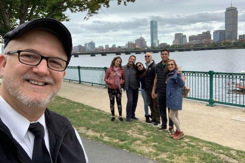 En grupp människor står på en strandpromenad vid en flod, med en stadsbild i bakgrunden. 2