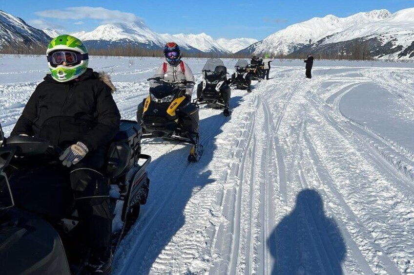 A group of people on snowmobiles on a snowy path with mountains in the background. 5