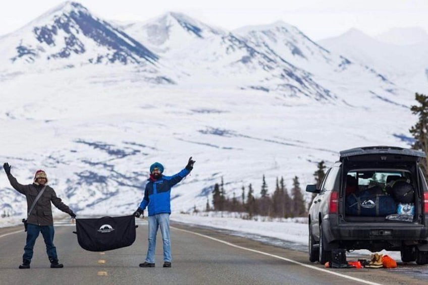Two people standing on a road with a car in the background and snow-covered mountains in the distance. 3