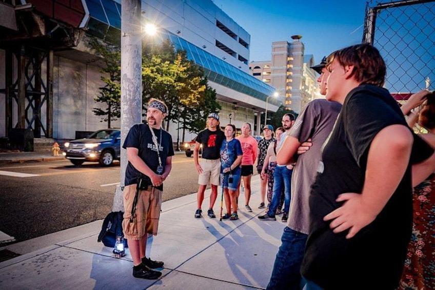 A group of people standing on a sidewalk at dusk, with buildings and a street in the background. 1
