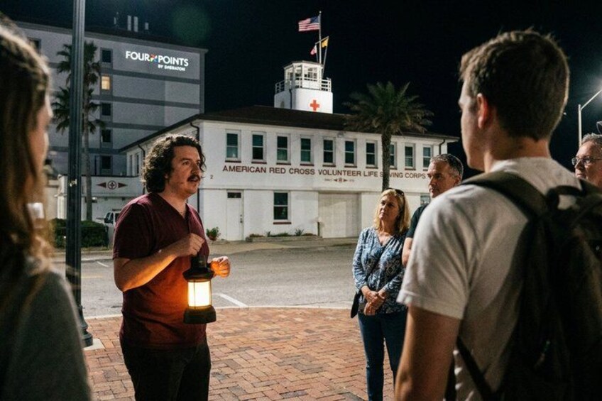 A group of people standing on a sidewalk at night, a building with a sign for the American Red Cross Volunteer Life Saving Corps, and a Four Points by Sheraton hotel in the background. 1