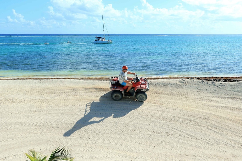 Una persona conduce un cuatrimoto sobre una playa arenosa, con un velero al fondo. 1