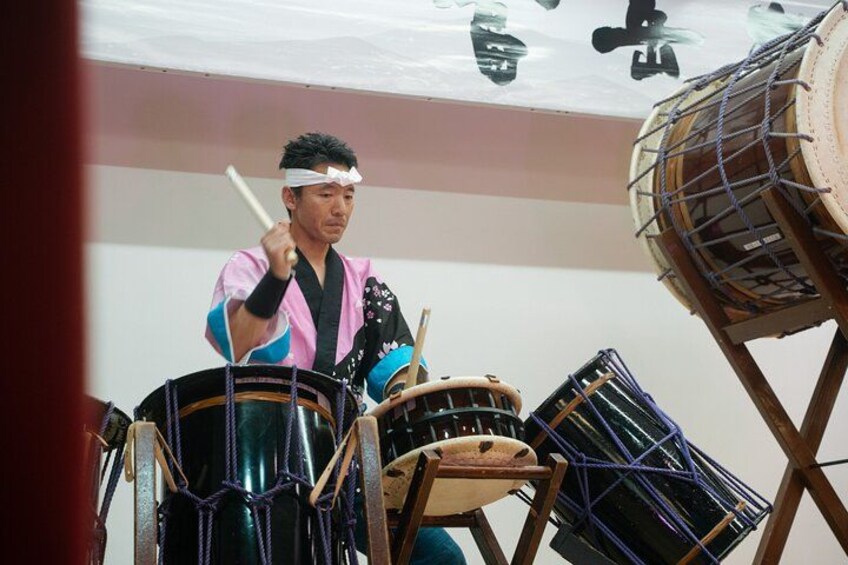 Eine Person spielt traditionelle Trommeln in einem Raum mit japanischer Kalligraphie an der Wand. 2