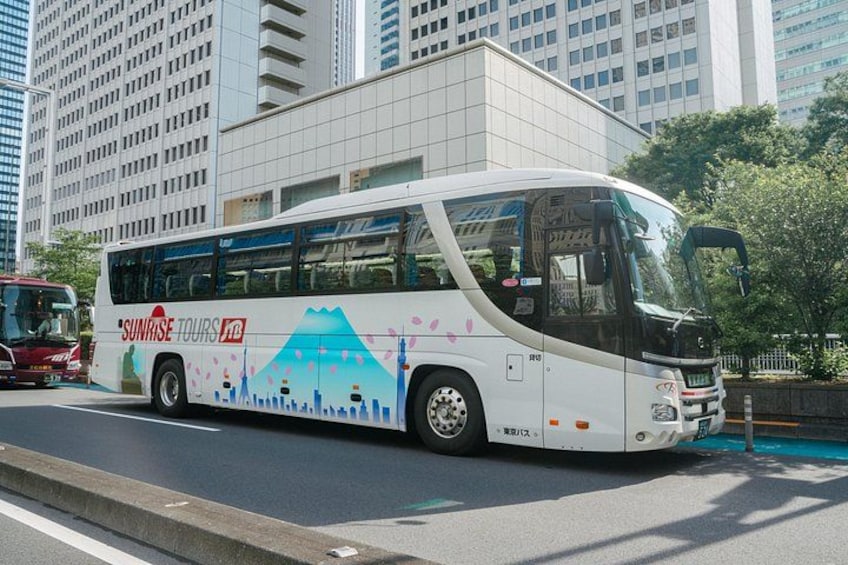 A white bus with 'Sunrise Tours' branding and a mountain design on its side, parked on a city street with modern buildings in the background. 3