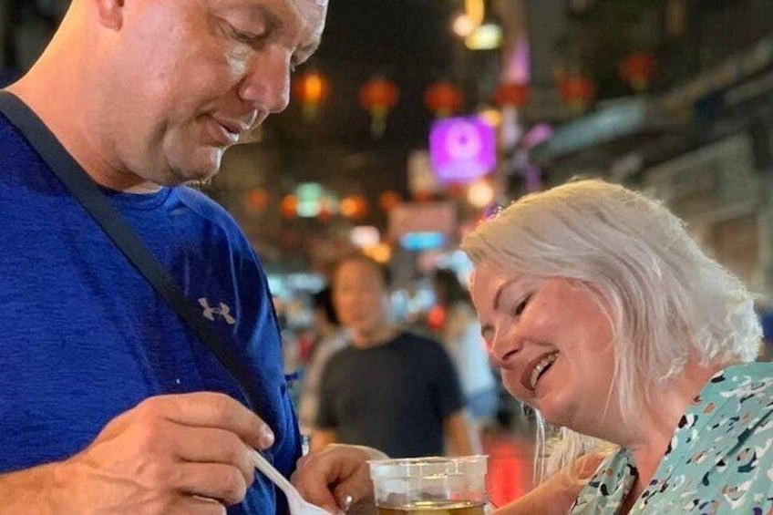 Dos personas comiendo en un puesto de comida callejera, con un fondo de calle concurrida. 3