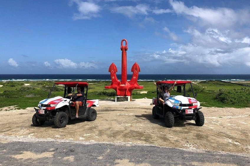 Two off-road vehicles parked on a dirt road with a large red anchor sculpture in the background. 1