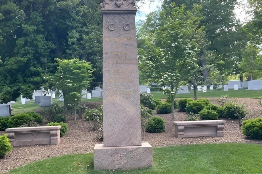 Een stenen monument met inscripties, omringd door een begraafplaats met grafstenen en vorm gesnoeide struiken. 2