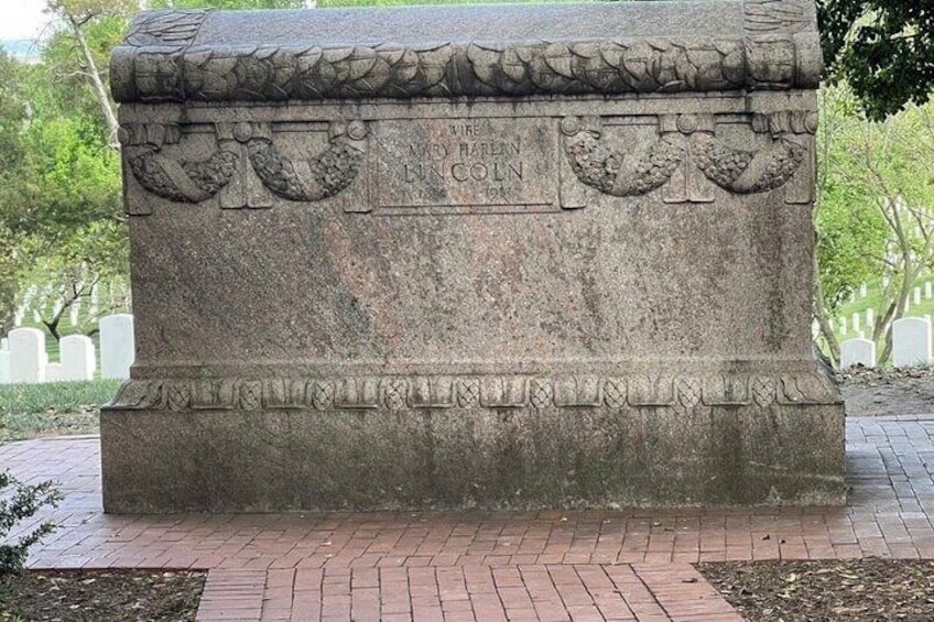 Een stenen monument met inscripties, gelegen op een met baksteen bestraat gebied, met bomen op de achtergrond. 3