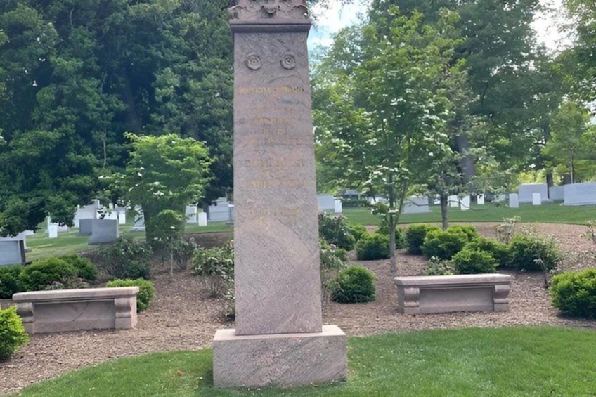 Ett stenmonument med inskriptioner, omgivet av en kyrkogård med vita gravstenar och välklippta buskar. 2
