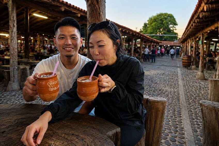 Two people sitting at a wooden table, holding clay pots with straws. 4