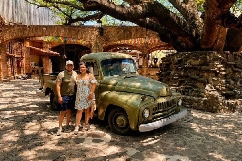 Un camion d’époque garé sous un grand arbre, un bâtiment rustique à la façade en briques et un sol pavé. 3