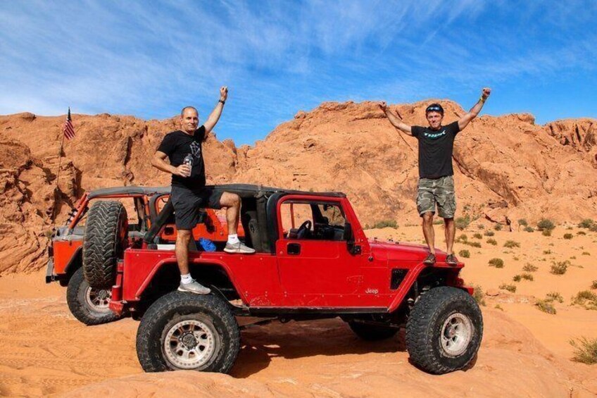 Two people standing on a red off-road vehicle in a desert landscape with red rock formations. 1