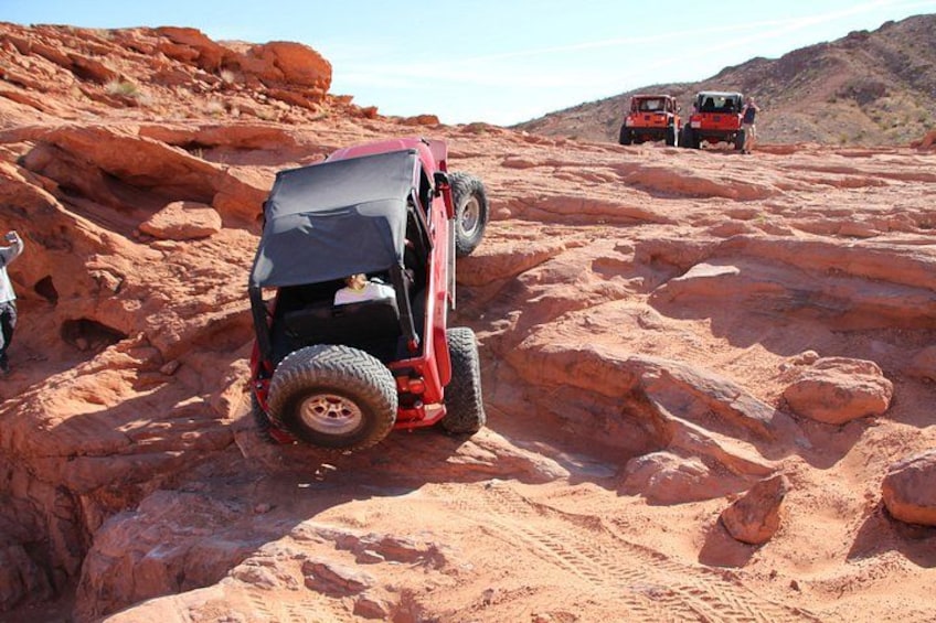 A red off-road vehicle stuck in a rocky terrain with another vehicle in the background. 2