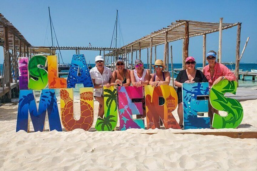 People standing in front of colorful letters spelling 'ISLANDS MUGORES' on a beach with a wooden structure in the background. 5