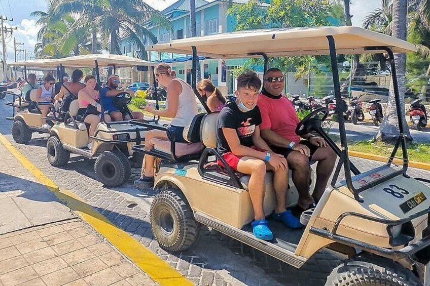 A group of people on a golf cart driving down a street with palm trees and buildings in the background. 4
