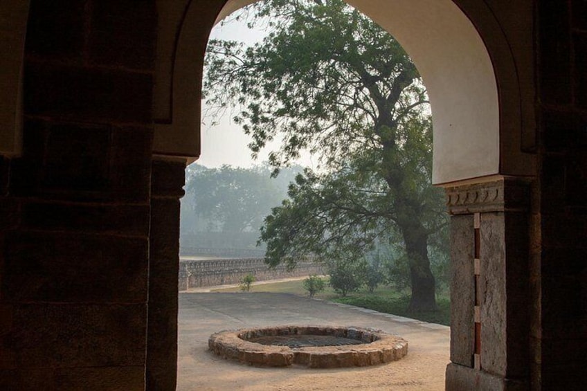 A stone archway leading to a circular stone structure, surrounded by trees and a distant bridge. 4
