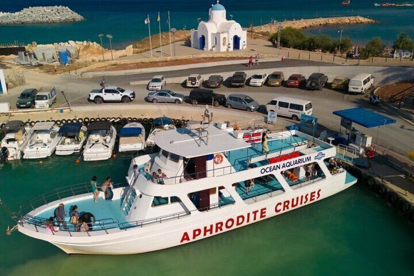 A white boat named 'Aphrodite Cruises' docked at a marina with several smaller boats and a building in the background. 3
