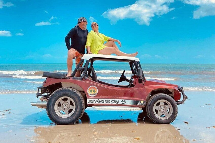 Een rood terreinvoertuig op een zandstrand met een helderblauwe lucht en de oceaan op de achtergrond. 1